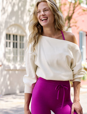 A woman outdoors wearing a cream off-the-shoulder sweatshirt and bright magenta high-waisted leggings with a drawstring waist.