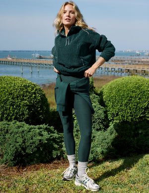 A woman stands outdoors near a waterfront and greenery, wearing a dark green fleece half-zip pullover, matching dark green leggings, white socks, and white sneakers.