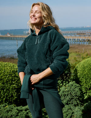 A woman outdoors wearing a dark forest green fleece pullover and matching green leggings, standing in front of green hedges with a pier and water in the background.