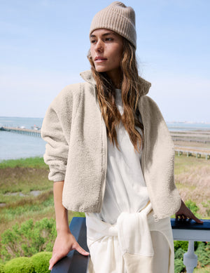A woman wearing a beige sherpa zip-up jacket, cream tie-front dress, and beige knit beanie stands outdoors by a railing with water and grass in the background.