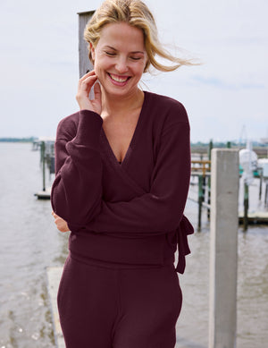 A woman smiling outdoors near a waterfront dock, wearing a matching burgundy knit wrap top and pants set.