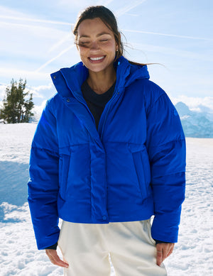 A woman smiling in a bright blue puffer jacket, standing in a snowy outdoor mountain setting.