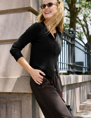 A woman wearing a black long-sleeve button-up shirt and dark brown wide-leg pants, posing outdoors in front of a stone wall and black metal fence.