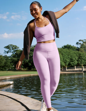 Woman wearing a purple racerback cropped tank and purple high-waisted midi leggings walking in front of a water feature.