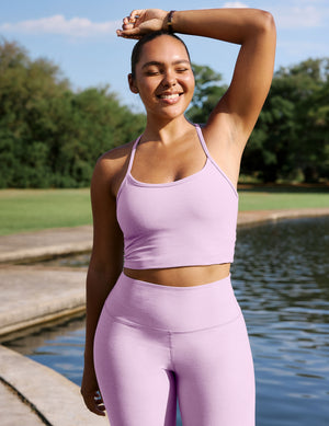 A woman wearing a light purple/lavender athletic crop top and matching high-waisted leggings stands outdoors by a lake with trees in the background.