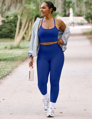 A woman walking outdoors on a park path wearing a royal blue sports bra and matching high-waisted leggings, with a light gray shirt draped off her shoulders and white sneakers.
