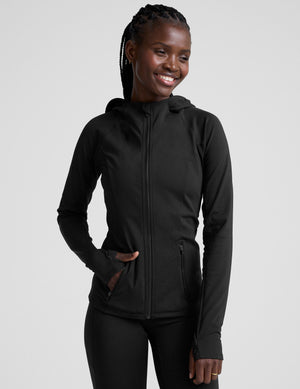 woman in black zip-up hooded jacket and leggings against a plain background. 