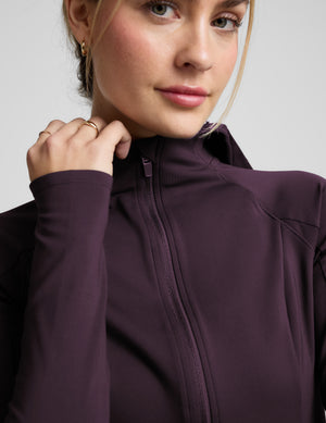 Woman wearing a purple athletic jacket and leggings on a light gray background. 