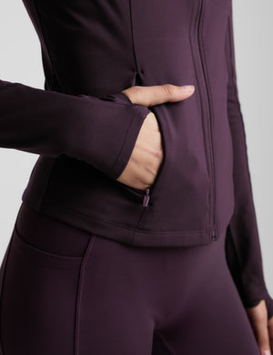 Woman wearing a purple athletic jacket and leggings on a light gray background. 