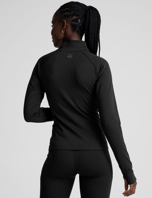 woman wearing a black long-sleeve athletic jacket with a zipper on a plain background. 