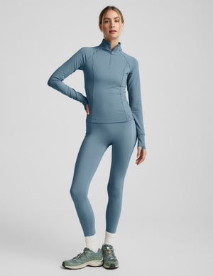 Woman wearing a blue quarter zip pullover and blue leggings on a light gray background. 