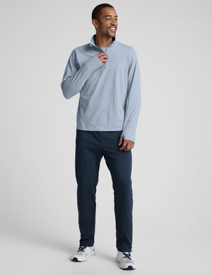 Man wearing a light blue quarter zip pullover and blue pants on a plain background. 