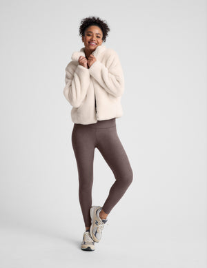 model wearing a light faux fur zip-up jacket and brown leggings against a plain background. 
