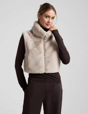 Woman wearing a beige faux fur vest over a dark outfit on a light gray background. 