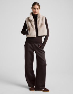 Woman wearing a beige faux fur vest over a dark outfit on a light gray background. 