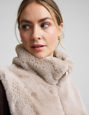 Woman wearing a beige faux fur vest over a dark outfit on a light gray background. 