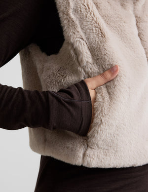 Woman wearing a beige faux fur vest over a dark outfit on a light gray background. 