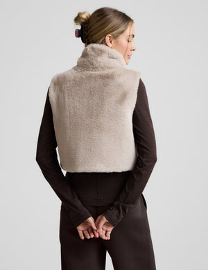 Woman wearing a beige faux fur vest over a dark outfit on a light gray background. 