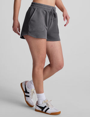 Model walking in dark gray Cali Short with a drawstring elastic waist, patch pockets, and curved dolphin hem.