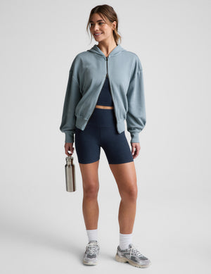 Woman wearing a blue zip-up jacket and blue biker shorts against a plain background. 