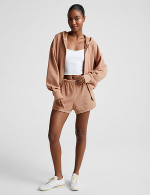 Woman wearing a brown zip-up jacket, white tank, and brown fleece shorts against a plain background. 