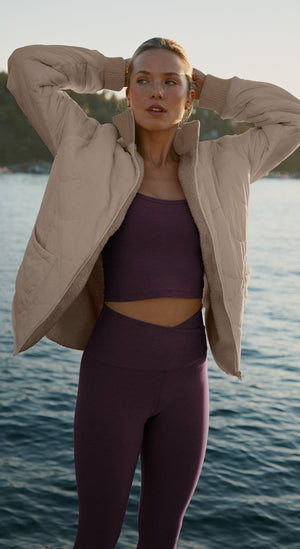 woman in a beige jacket and purple athletic wear standing by a body of water.