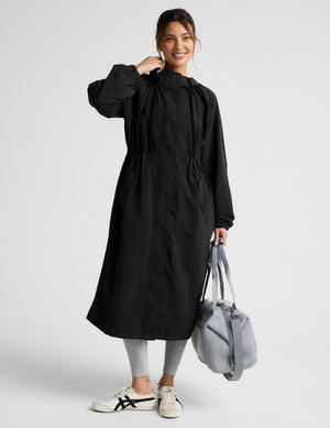 Woman wearing a black trench coat over a gray sports bra and leggings on a plain background. 