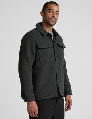 man wearing a dark gray shirt jacket over a gray shirt on a plain background. 