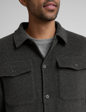 man wearing a dark gray shirt jacket over a gray shirt on a plain background. 