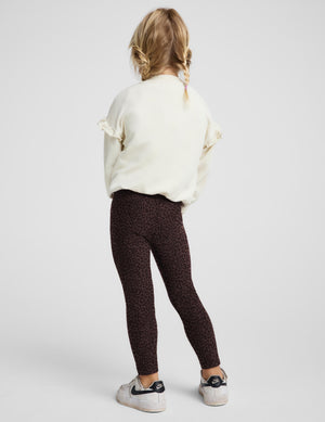 A young girl posing in dark brown leopard-print leggings, a cream puff-sleeve sweatshirt, and white sneakers against a light gray studio background.