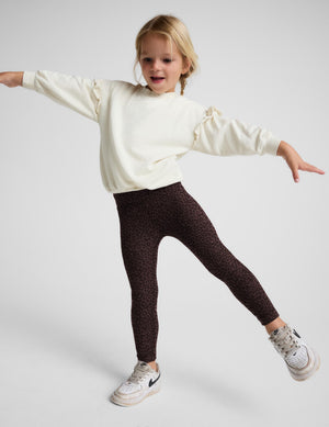 A young girl posing in dark brown leopard-print leggings, a cream puff-sleeve sweatshirt, and white sneakers against a light gray studio background.