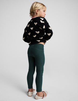 A young girl with blonde hair is shown from the back wearing a black knit sweater with a cream heart pattern and dark green leggings, standing against a light gray studio background.