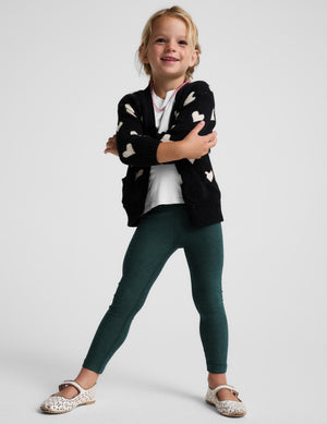 A young girl modeling a black knit cardigan with cream hearts, dark green leggings, a white t-shirt, and white Mary Jane shoes, posed against a light gray studio background.