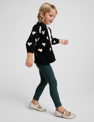 A young girl walking in profile wearing a black knit cardigan with white heart patterns, dark green leggings, and white Mary Jane shoes with a black pattern, against a light gray studio background.