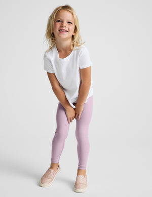 A young girl with blonde hair smiling and looking up, wearing a white t-shirt, light purple leggings, and pink slip-on shoes, photographed against a solid light gray background.