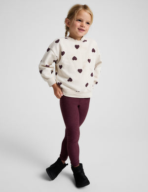 A young girl modeling a cream-colored fleece hoodie with a dark burgundy heart pattern, paired with burgundy leggings and black boots, against a light gray studio background.