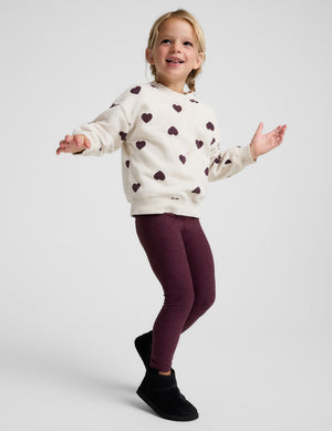 A young girl modeling a cream-colored fleece hoodie with a dark burgundy heart pattern, paired with burgundy leggings and black boots, against a light gray studio background.