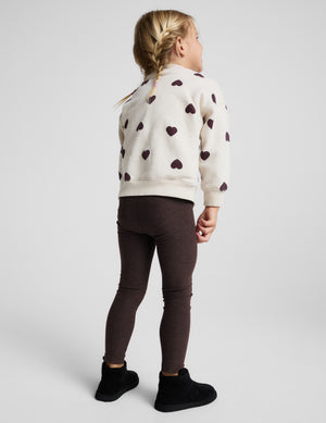 A young girl stands against a light gray studio background wearing a cream knit sweatshirt with dark brown heart patterns, dark brown leggings, and black ankle boots.