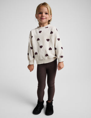 A young girl stands against a light gray studio background wearing a cream knit sweatshirt with dark brown heart patterns, dark brown leggings, and black ankle boots.