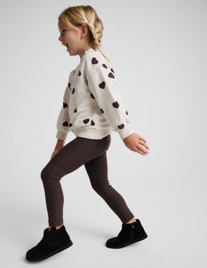 A young girl stands against a light gray studio background wearing a cream knit sweatshirt with dark brown heart patterns, dark brown leggings, and black ankle boots.