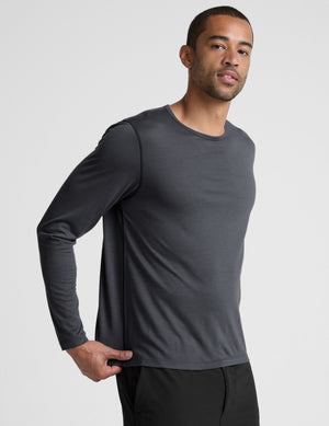 Man wearing a dark gray crew neck long sleeve and black pants against a light background. 