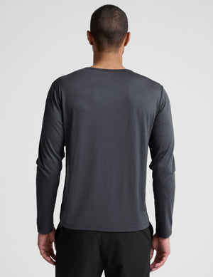Man wearing a dark gray crew neck long sleeve and black pants against a light background. 