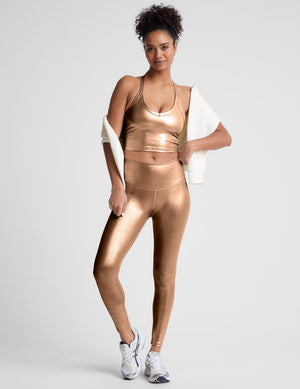 woman wearing a gold v-neck cropped tank and leggings on a plain background. 