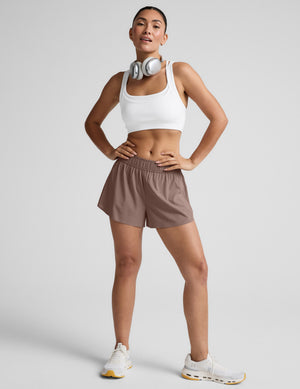 Woman wearing a white sports bra and brown shorts on a plain background. 