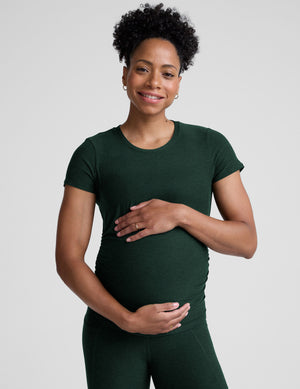 model woman wearing a dark green maternity t-shirt and leggings against a light gray background. 