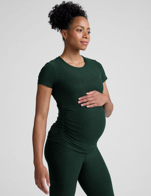 model woman wearing a dark green maternity t-shirt and leggings against a light gray background. 