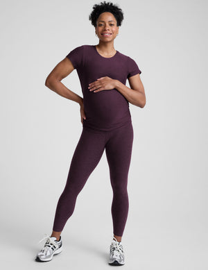 model wearing a purple maternity t-shirt and leggings against a light gray background. 