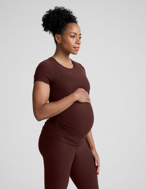 Pregnant woman wearing a brown maternity t-shirt and leggings against a plain background. 