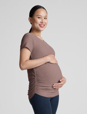 Pregnant woman wearing a brown shirt and blue leggings against a plain background. 