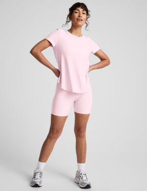 Woman wearing a pink short sleeve top and biker shorts on a light gray background. 